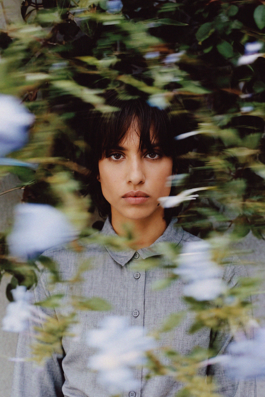 A woman with dark hair and bangs wearing a light gray button-up shirt, framed by soft-focus green leaves and pale blue flowers in the foreground.