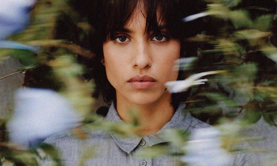 A woman with dark hair and bangs wearing a light gray button-up shirt, framed by soft-focus green leaves and pale blue flowers in the foreground.