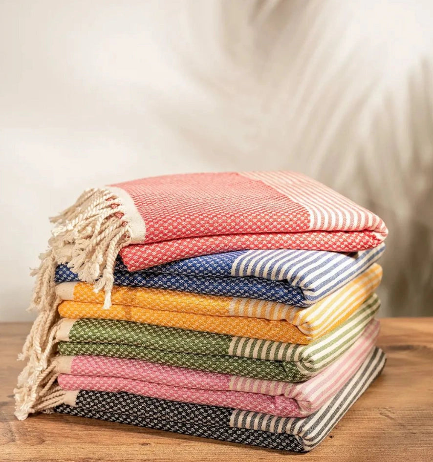 A stack of folded woven towels or blankets with fringed edges, shown on a wooden surface against a softly blurred neutral background. The stack includes multiple colors and stripe patterns: coral pink, blue, mustard yellow, olive green, light pink, navy, and cream.