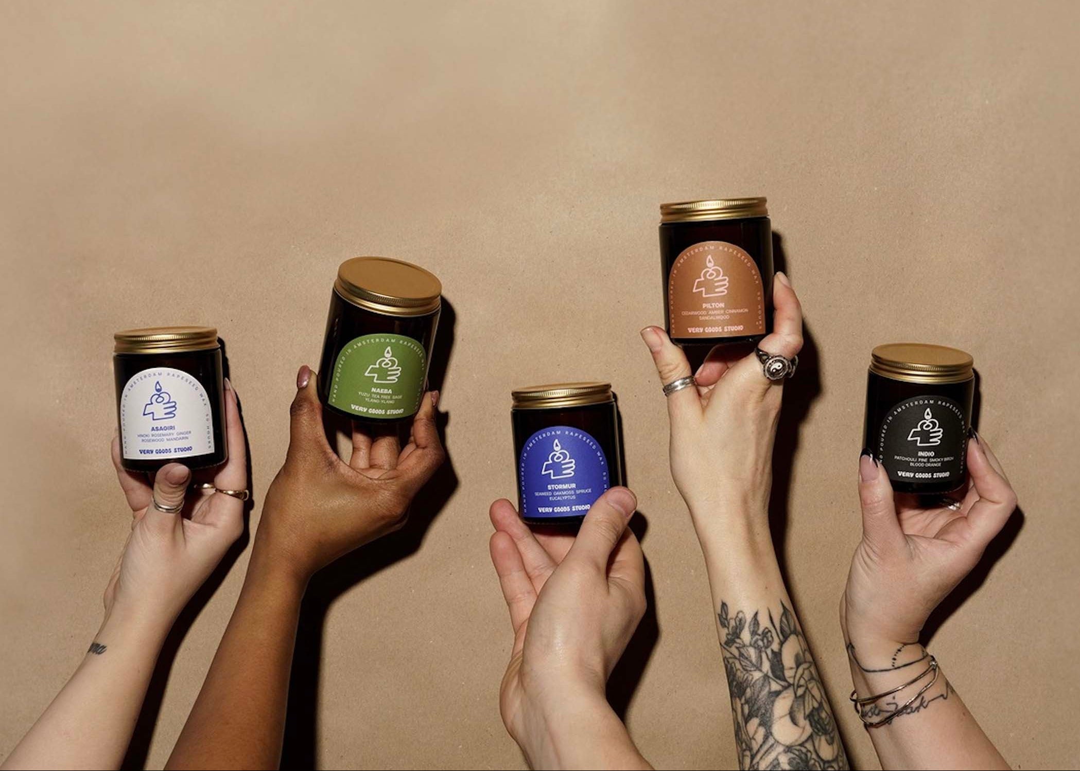 Five hands holding candles with visible labels against a beige background VERY GOOD STUDIO