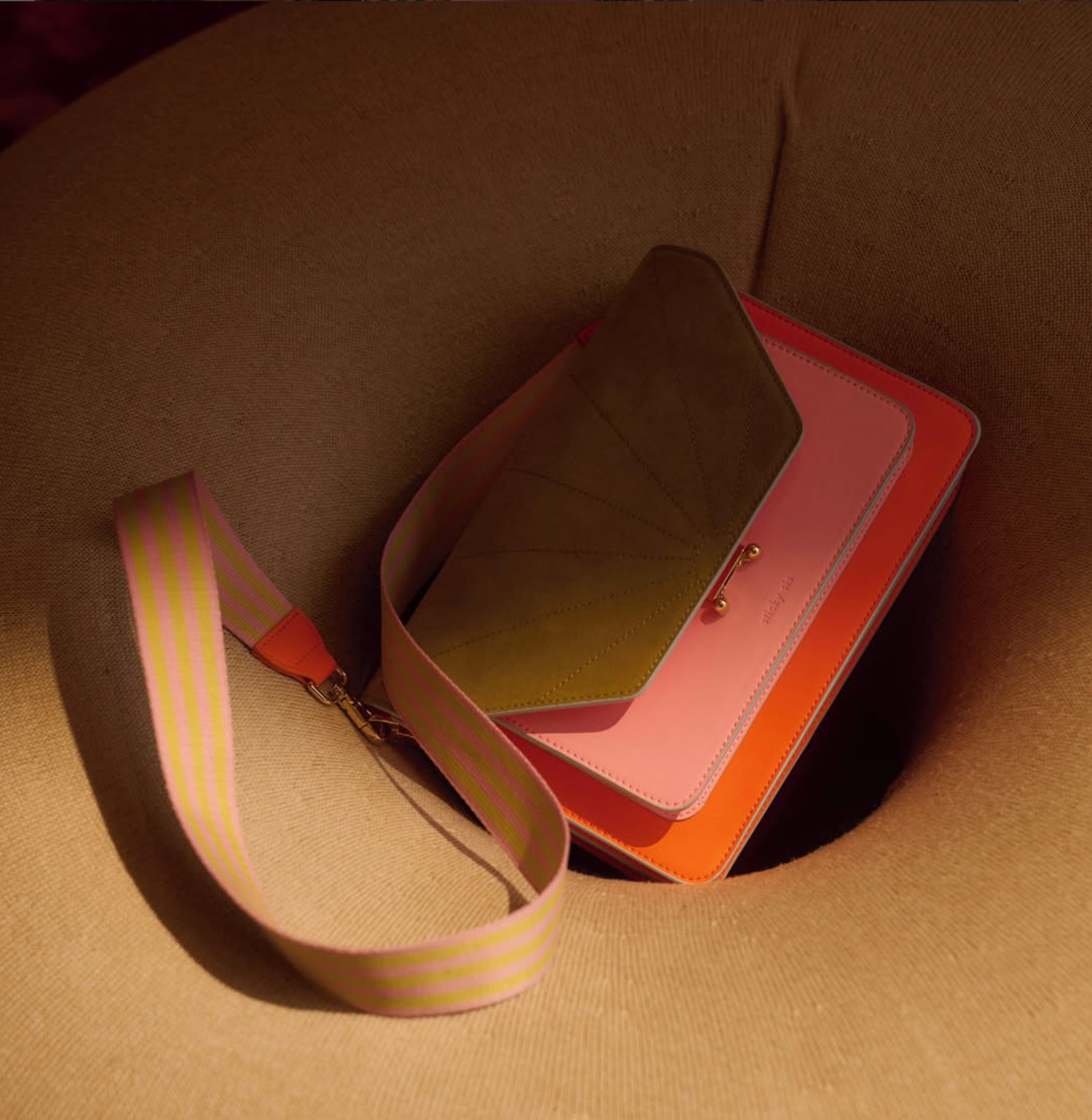 A structured handbag with a wide pink and yellow striped strap, resting inside a beige woven chair. The bag features a layered design with a dark olive-green top flap, a pink body, and an orange base, accented by gold-tone hardware.
