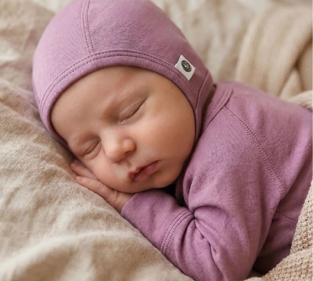A sleeping newborn baby wearing a dusty purple knit bonnet and matching long-sleeve top, resting on a textured beige blanket.